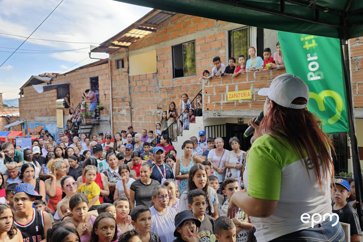 Fotografía de una funcionaria de EPM dirigiéndose a la comunidad con un micrófono, mientras al fondo se observa una multitud participando activamente en el encuentro.