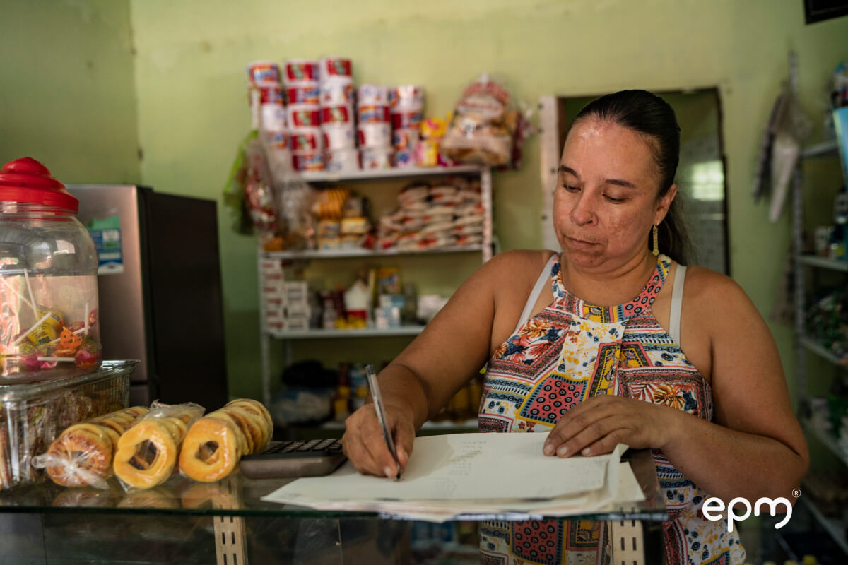 Fotografía de una mujer tomando notas en una hoja de papel, de fondo se puede percibir una tienda de productos 