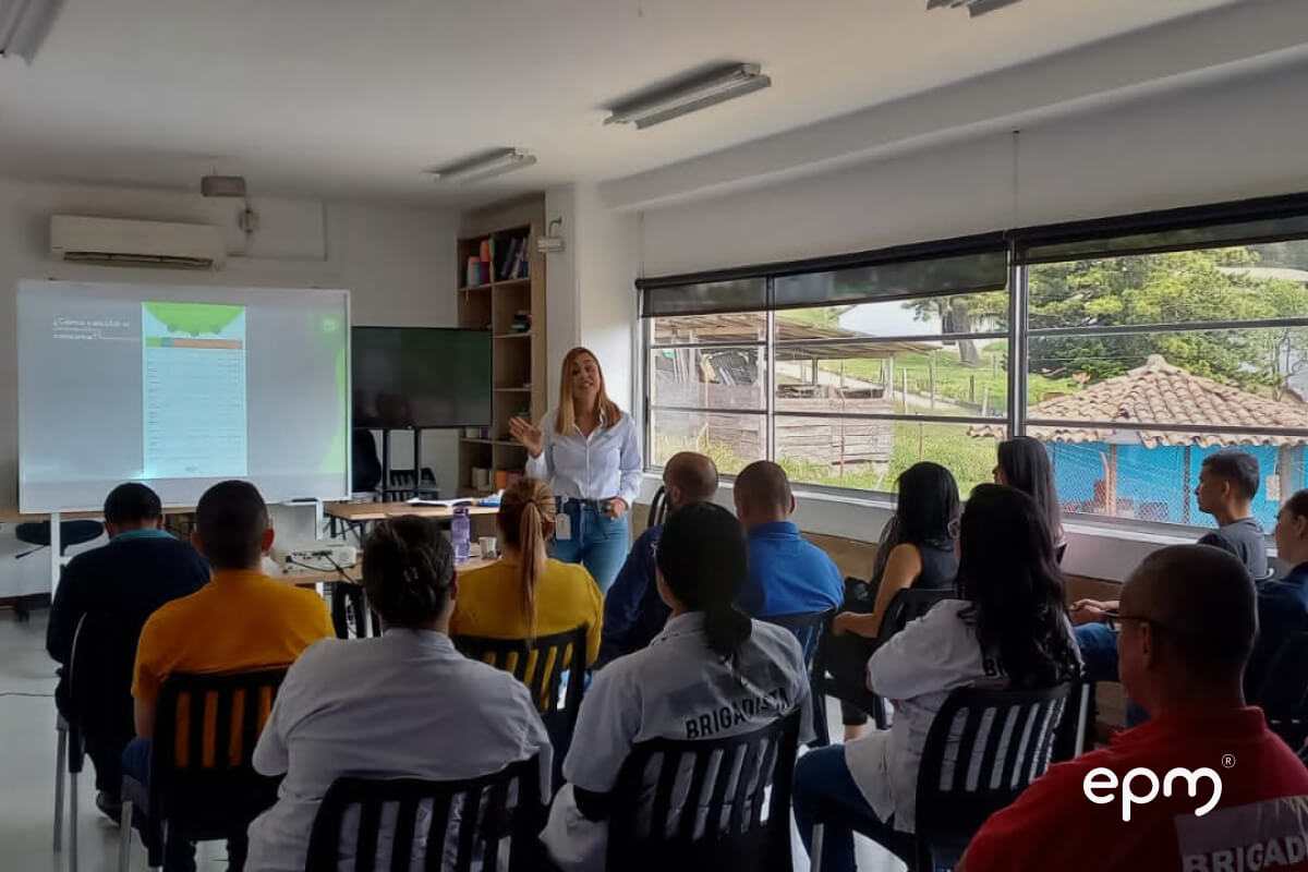 Fotografía de una mujer de pie dando una charla a un grupo de personas sentadas en un aula. En la pantalla se proyecta información educativa relacionada con EPM, mientras la luz natural entra por amplias ventanas que muestran un paisaje rural.