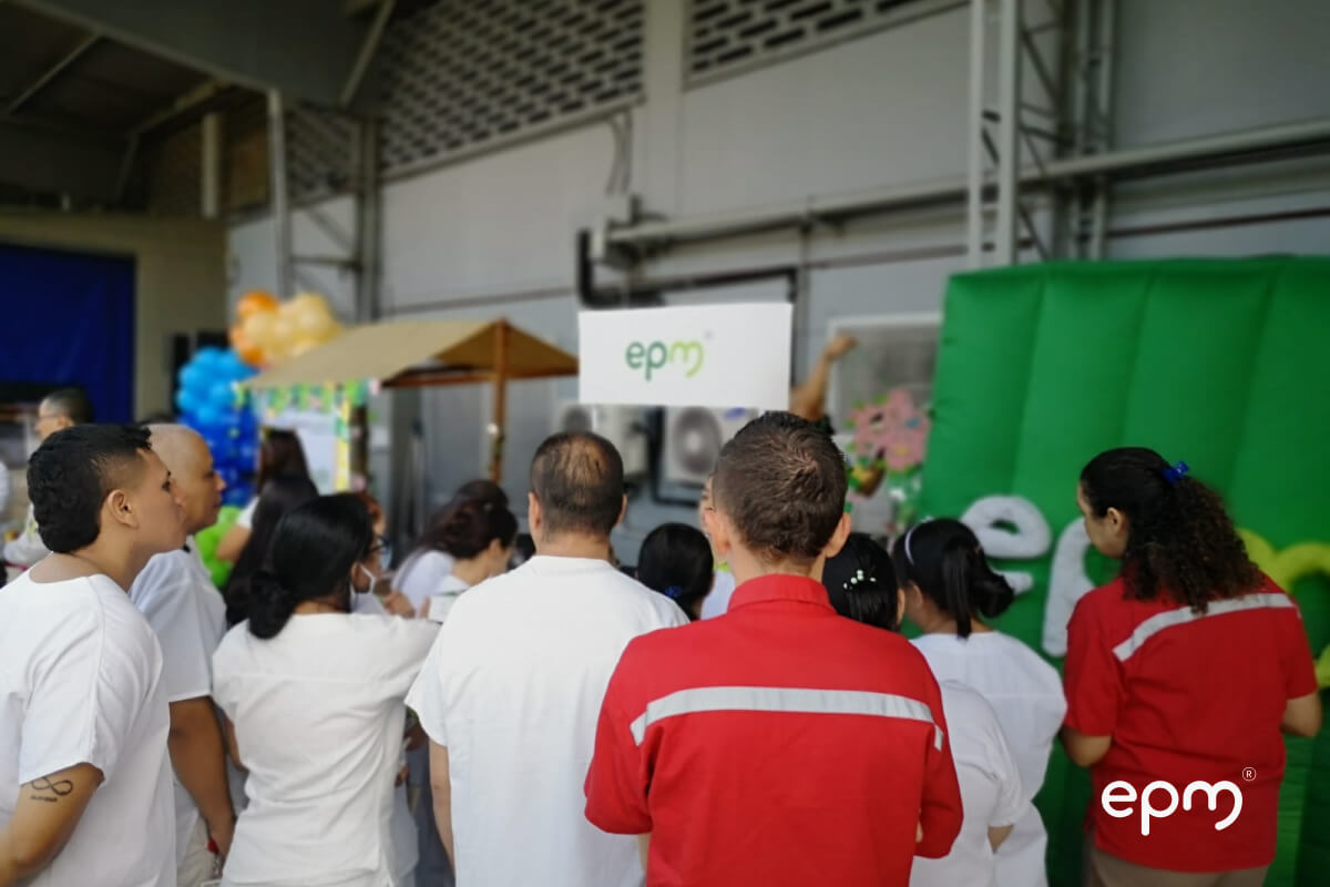 Fotografía de un grupo de trabajadores de EPM, realizando una actividad en un espacio cerrado. Al fondo, se ve un cartel con el logo de EPM y una estructura inflable verde como parte de la escenografía.