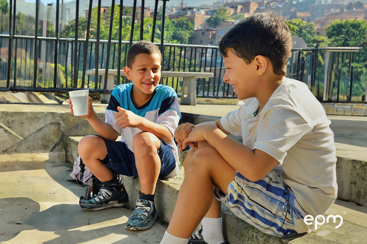 Niños sentados sonriendo bebiendo agua. Al fondo se observan casas, árboles y malla