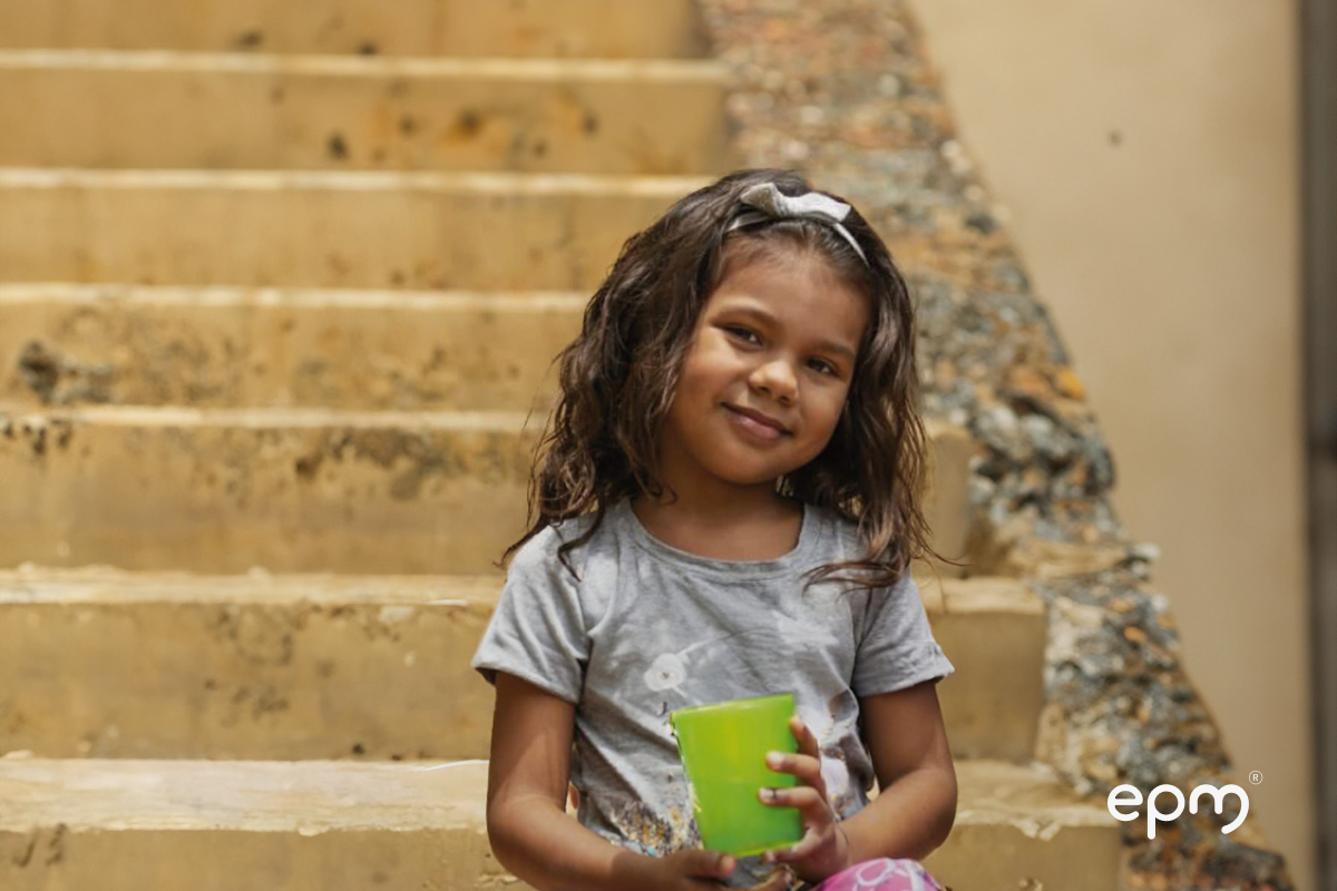 Niña con vaso verde en las manos sonriendo. De fondo se observan escalas en cemento