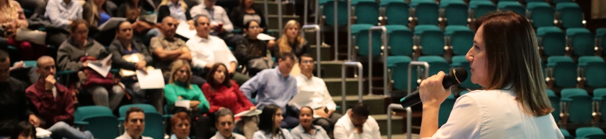 Asistencia Presencial. El acceso a está información tiene una imagen de una mujer con camisa blanca hablando por micrófono en un auditorio con personas sentadas escuchando