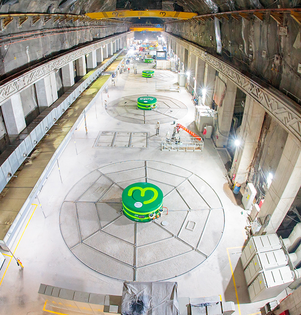 Vista panorámica de la casa de máquinas subterránea en Hidroituango, con varios generadores verdes alineados y personal técnico trabajando.