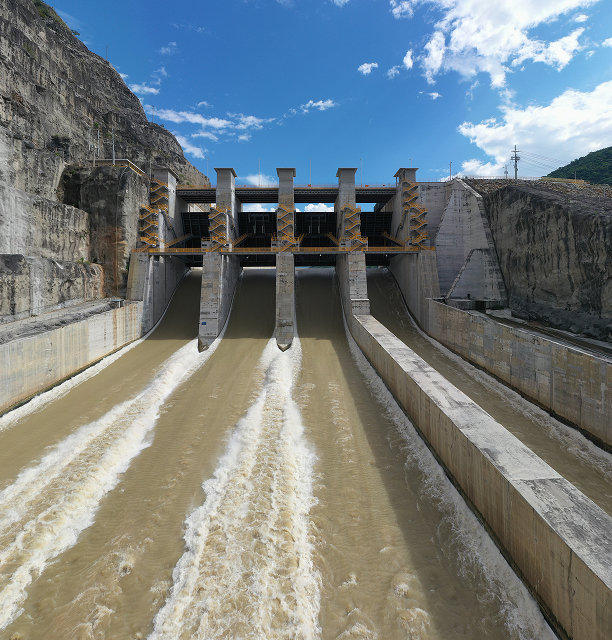 Imagen frontal del vertedero principal de la represa Hidroituango, con agua fluyendo por tres canales, enmarcado por las estructuras de contención de concreto.