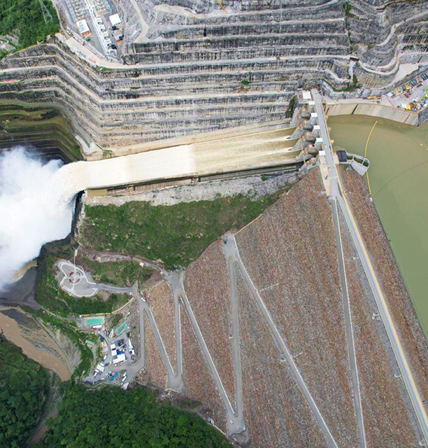 Fotografía aérea de la represa de Hidroituango mostrando la descarga de agua a través de los vertederos, rodeada por estructuras escalonadas y el embalse.