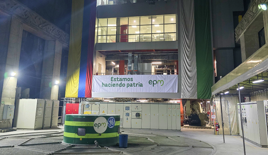 Vista del interior de la casa de máquinas de Hidroituango con banderas de Colombia y EPM colgadas al fondo. En el centro, un generador verde con el logo de EPM, y un cartel que dice "Estamos haciendo patria".