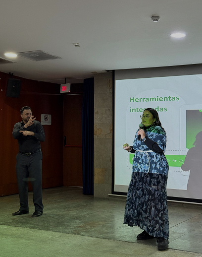 Mujer se dirige al público mientras es acompañada por un hombre que hace lenguaje de señas en una capacitación de accesibilidad