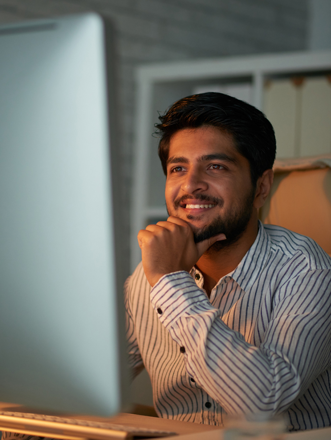Hombre sonriente con camisa de rayas, sentado frente a un computador de escritorio, mirando la pantalla con expresión de interés.