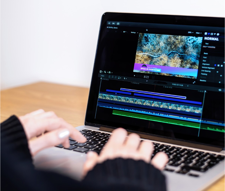 Manos de una persona trabajando en un computador portátil, editando un video en un software de edición con líneas de tiempo y pistas de audio y video visibles en la pantalla.