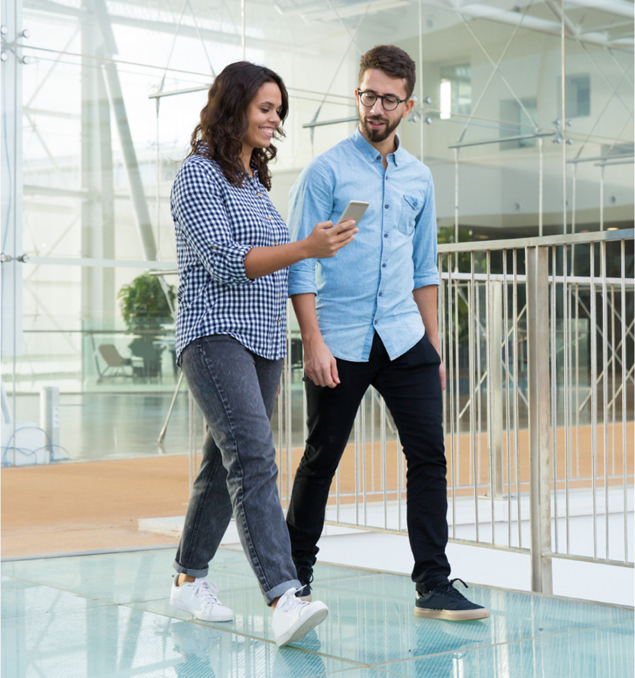 Mujer y hombre jóvenes caminando en un espacio moderno de vidrio y metal, mirando juntos la pantalla de un teléfono móvil con expresión de interés y conversación.