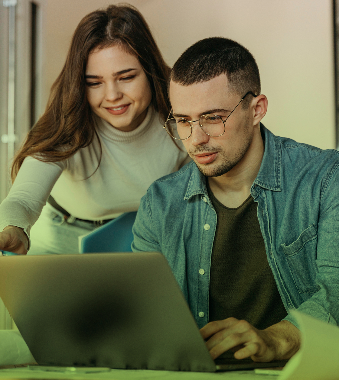 Dos personas trabajando en equipo frente a un computador portátil; una mujer sonríe y señala la pantalla mientras un hombre con gafas escribe en el teclado.