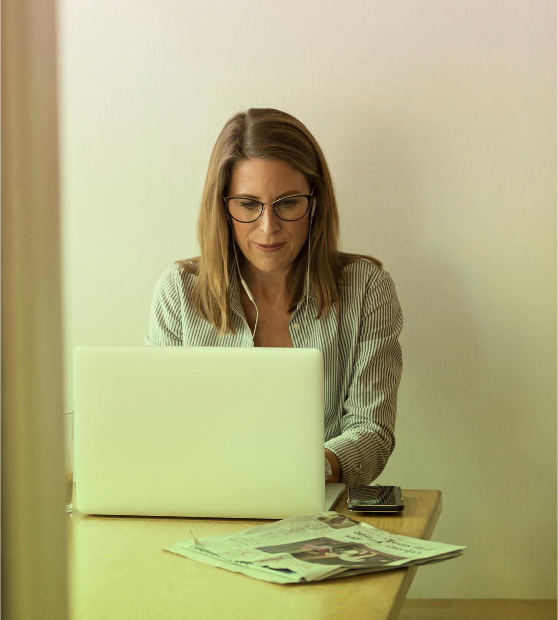 Mujer adulta con gafas y audífonos trabajando en un computador portátil, con un periódico y un celular sobre la mesa.