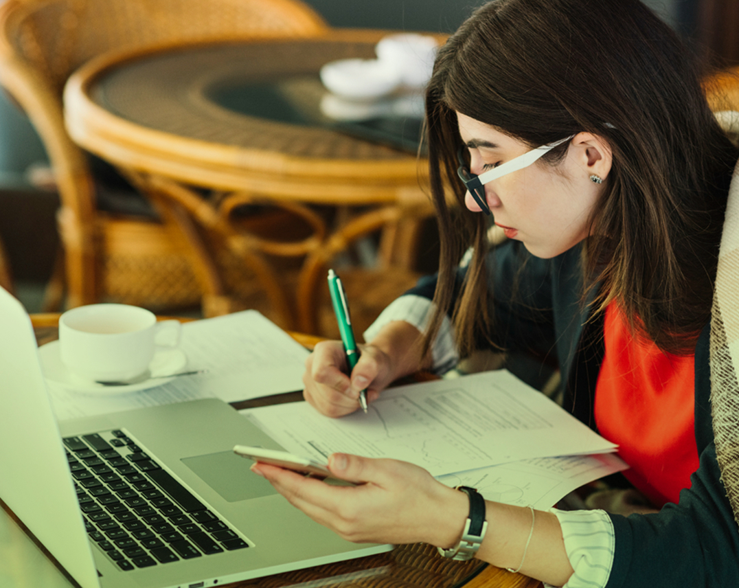 Mujer joven con gafas trabajando en una mesa, tomando notas en documentos impresos mientras sostiene un celular y tiene un computador portátil abierto al frente.