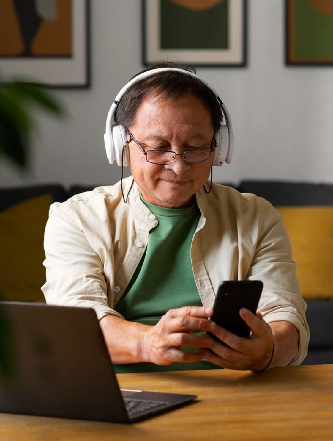 Hombre mayor con audífonos y gafas utiliza su celular mientras sonríe, sentado frente a un computador portátil en una sala.