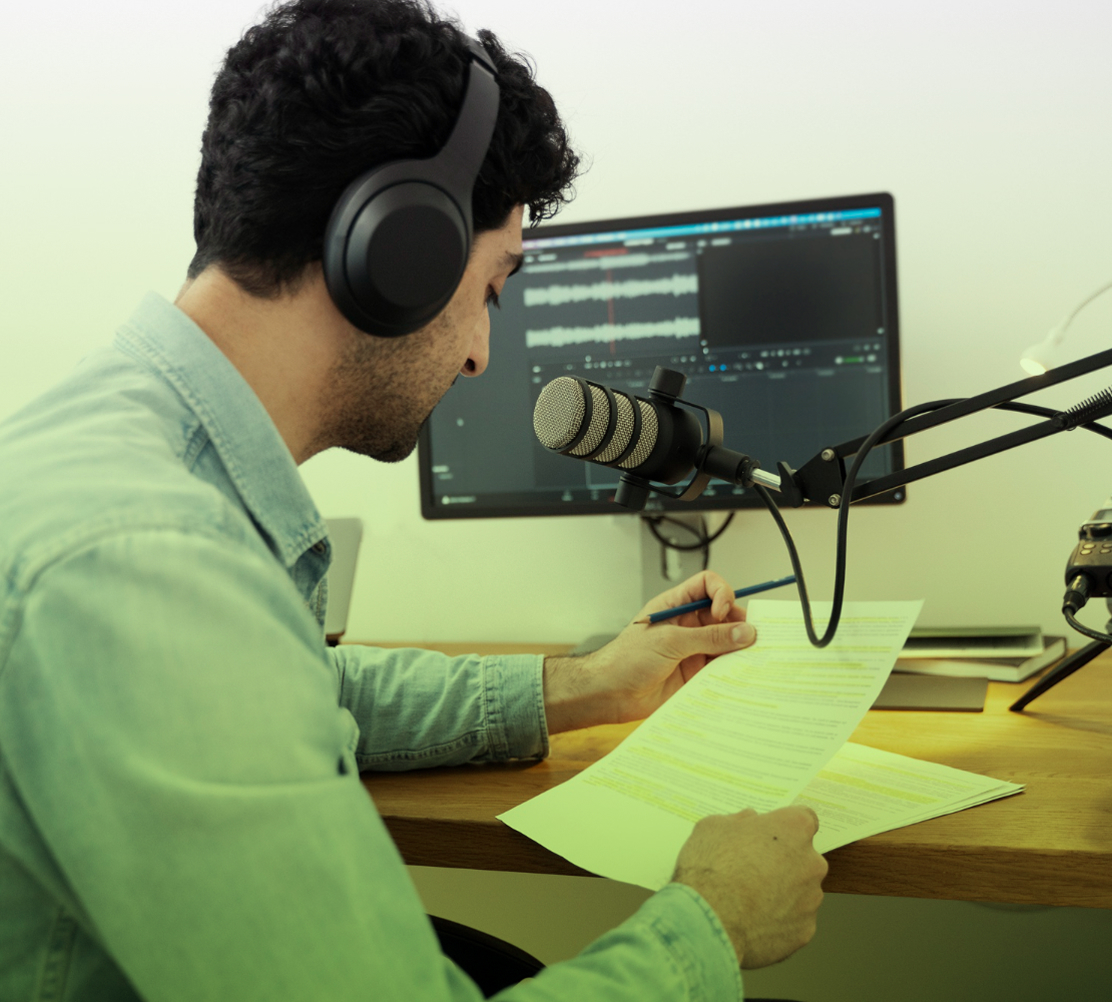 Hombre con audífonos frente a un micrófono de condensador, leyendo un documento mientras graba en un estudio casero con un computador en segundo plano.