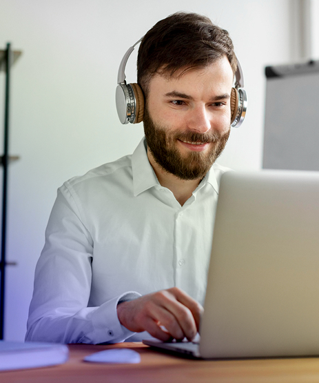 Hombre joven con audífonos participa en un curso virtual desde su computador portátil, sonriendo mientras trabaja.