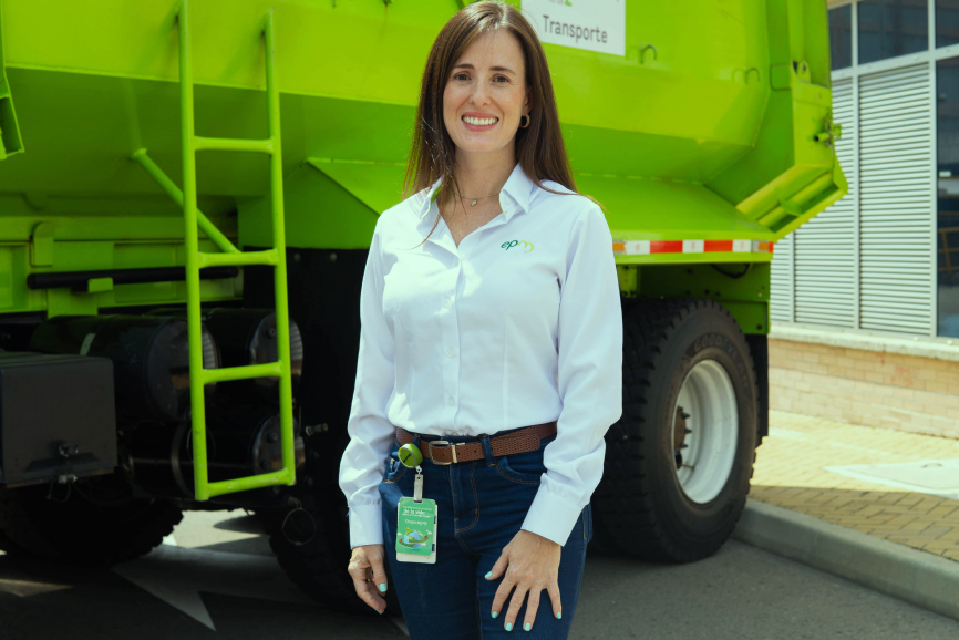 Mujer trabajadora de EPM con camisa blanca corporativa, sonriendo de pie frente a un vehículo de hidrógeno verde