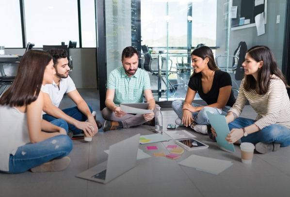 Grupo de jóvenes reunidos en el piso de una oficina moderna, trabajando en equipo con computadoras portátiles, documentos y notas adhesivas