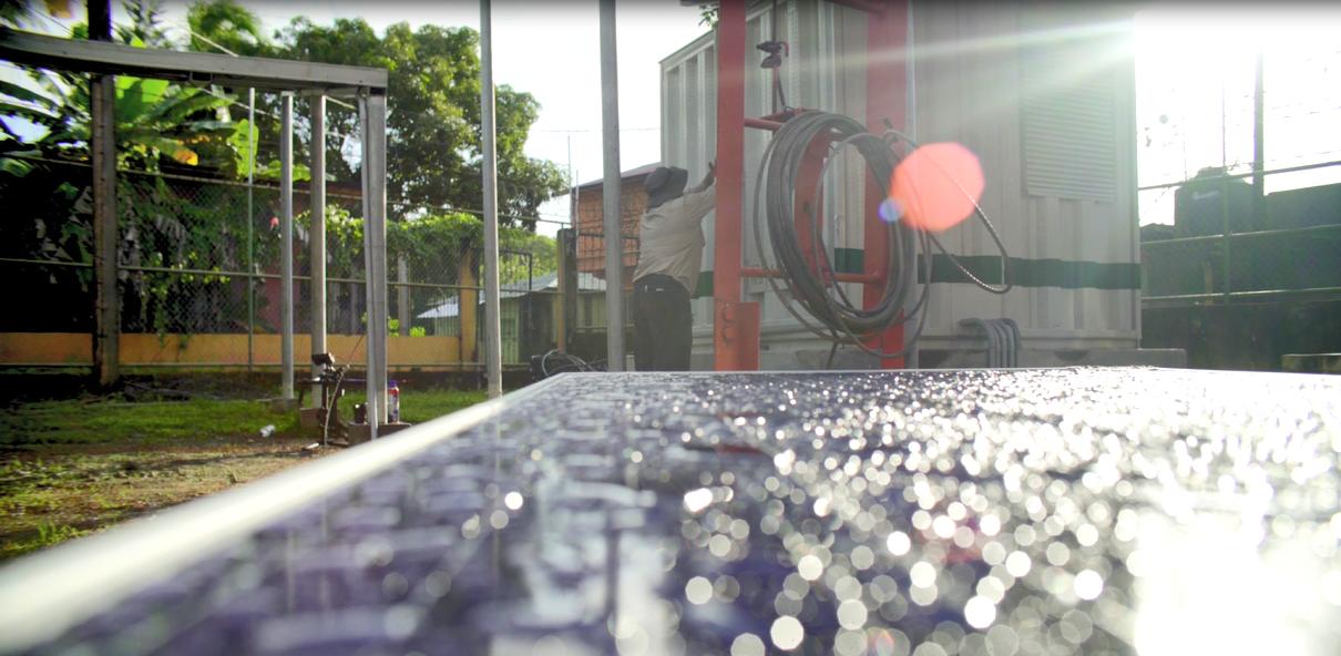 Técnico revisa unidad de generación eléctrica en contenedor mientras panel solar cubierto de rocío refleja la luz del sol