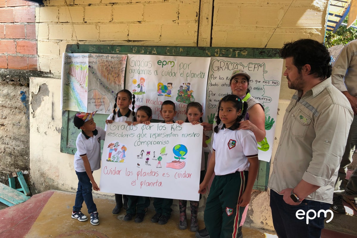 Niños presentando un cartelera que dice: "Gracias por los kits escolares que representan nuestro compromiso. Cuidar las plantas es cuidar el planeta". Atrás los acompañan dos funcionarios de EPM