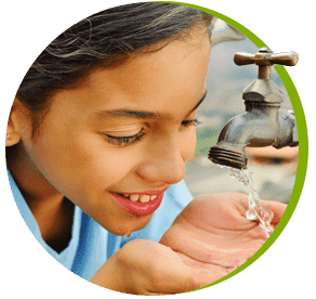 Niña sonriente bebiendo agua de un grifo con las manos en forma de cuenco.