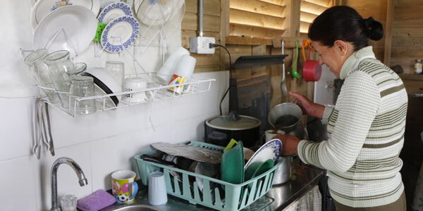 Mujer lavando utensilios en una cocina con estanterías y platos organizados.