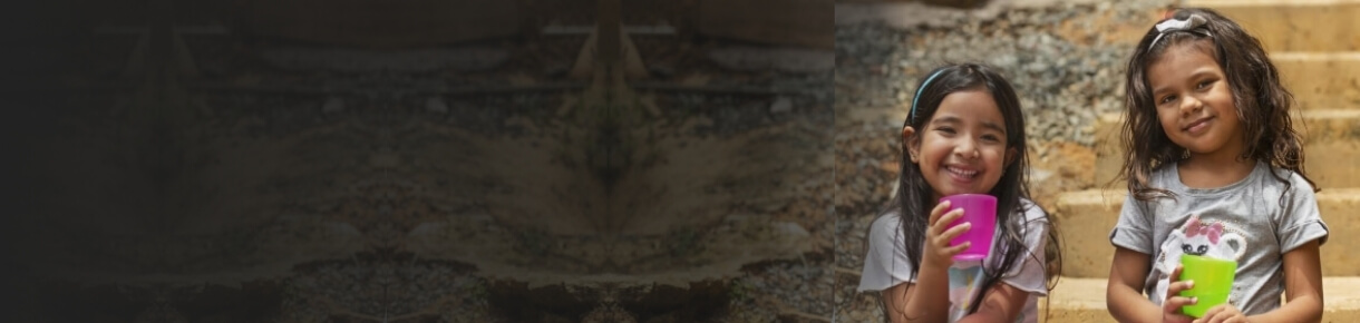 Dos niñas sonrientes sentadas en unas escaleras al aire libre, sosteniendo vasos de colores mientras disfrutan agua potable en una comunidad rural