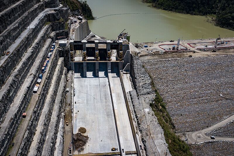 Vista aérea de la estructura principal de la represa Hidroituango, con vertederos secos, terrazas escalonadas en la ladera izquierda y el embalse de agua al fondo.