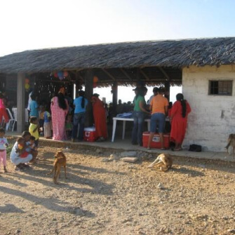 Personas en el parque eólico Jepírachi reunidas en una estructura con techo de palma en un entorno rural, algunas interactuando y otras observando, con niños y perros presentes