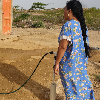 Mujer indígena Wayuu con vestido tradicional azul, extrayendo agua de una bomba manual en un entorno rural