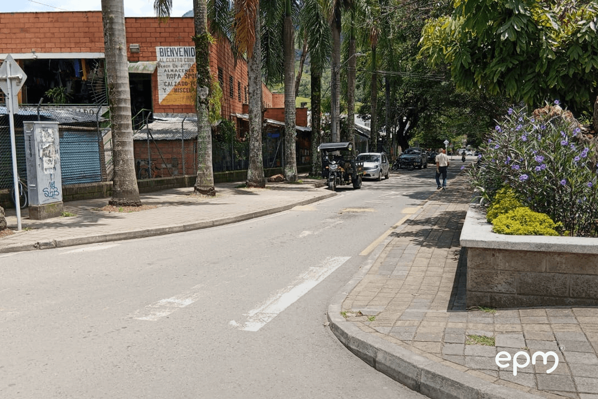 Fotografía de una calle tranquila bordeada por palmas altas, con edificios de ladrillo y un cartel que da la bienvenida a un centro comercial.
