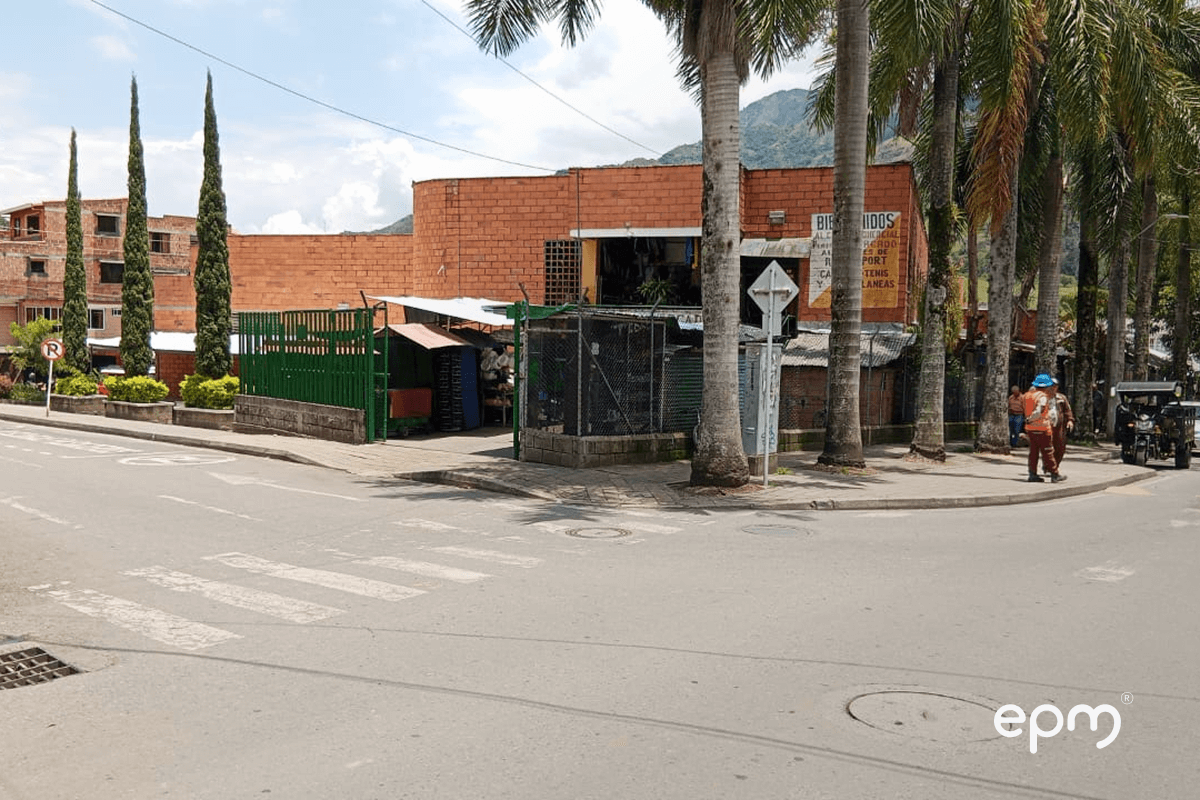 Fotografía de la vista exterior de un edificio de ladrillo con techo blanco en una esquina, rodeado de árboles altos y palmas.
