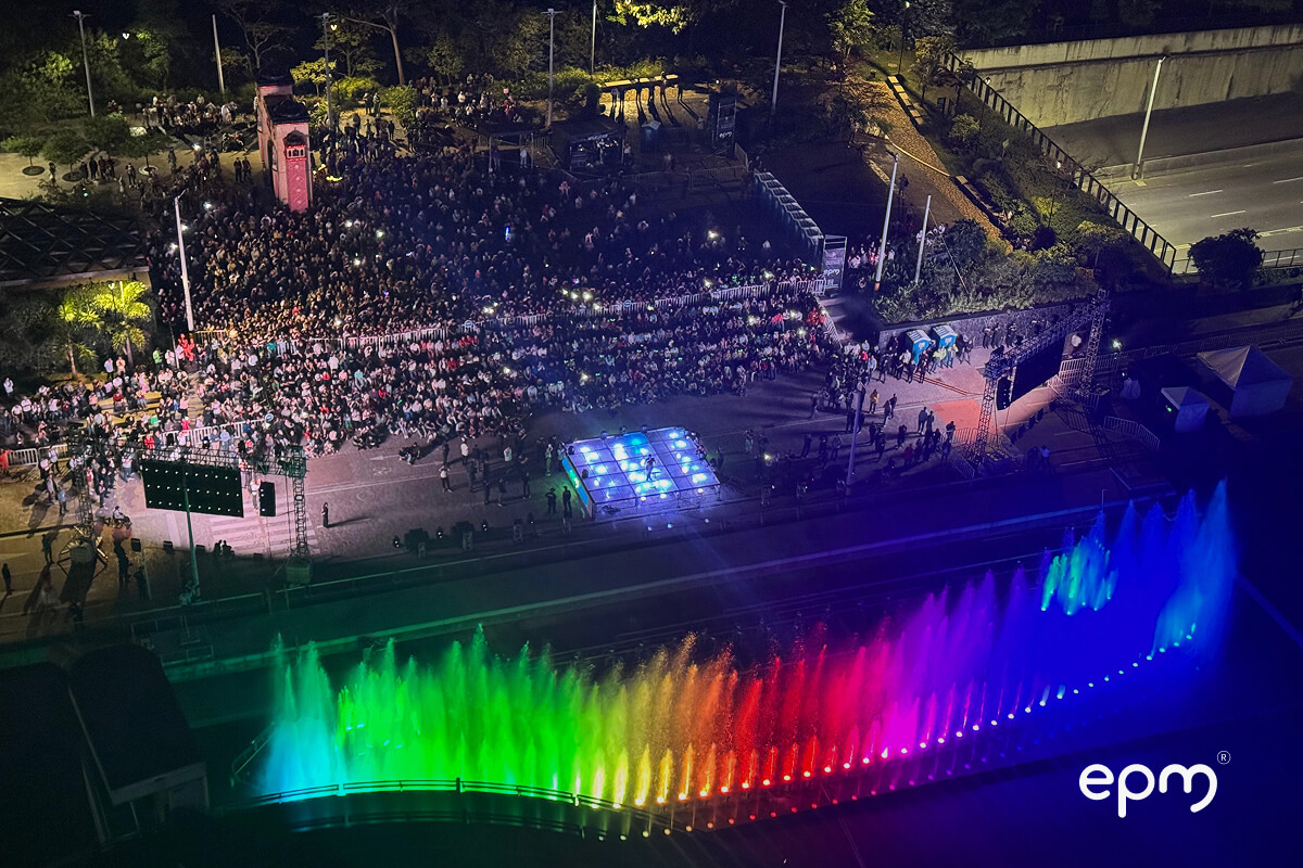 Vista aérea nocturna de una multitud disfrutando el encendido de los Alumbrados Navideños de EPM, con un escenario y fuente de luces arcoíris