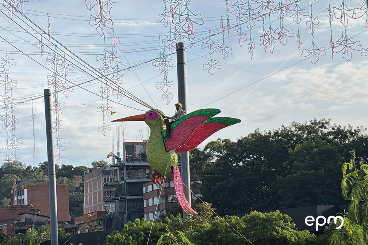 Figura de pájaro en color verde y rojo de alumbrados navideños de EPM. Al fondo se observan casas y árboles