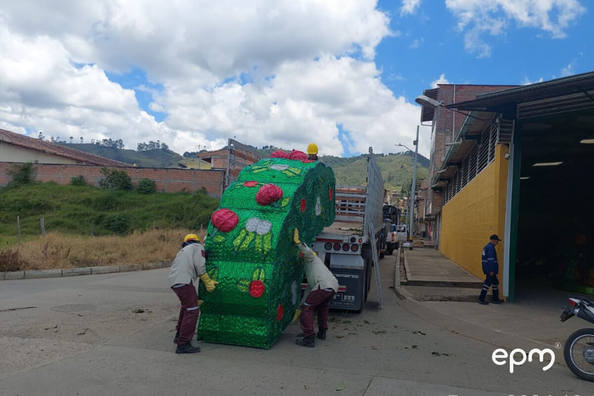 Trabajadores cargando una estructura decorativa verde en un camión para transporte, en un entorno urbano con cielo despejado