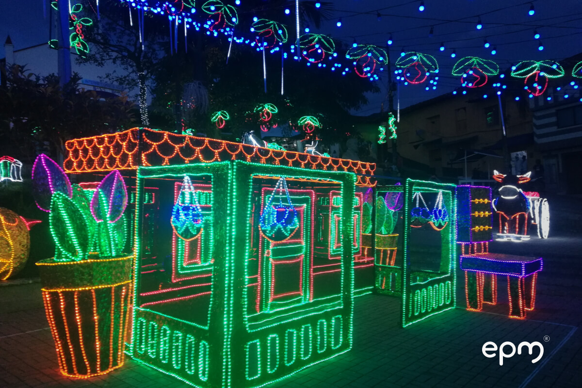 Decoración navideña iluminada con luces de colores en forma de casas y adornos colgantes en un espacio público nocturno