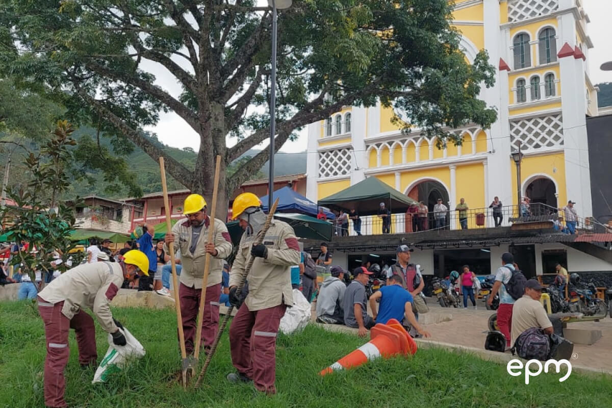 Trabajadores con cascos amarillos realizan labores de plantación en un parque frente a una iglesia colonial amarilla, con personas alrededor