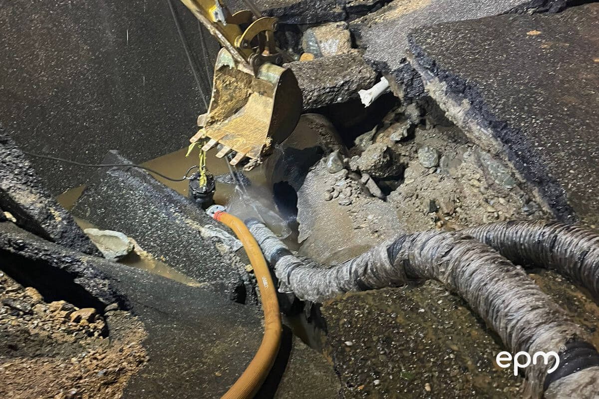 Excavadora realizando trabajos de reparación en una vía afectada por una fuga de agua, con tuberías expuestas y equipos de succión operando, durante la noche