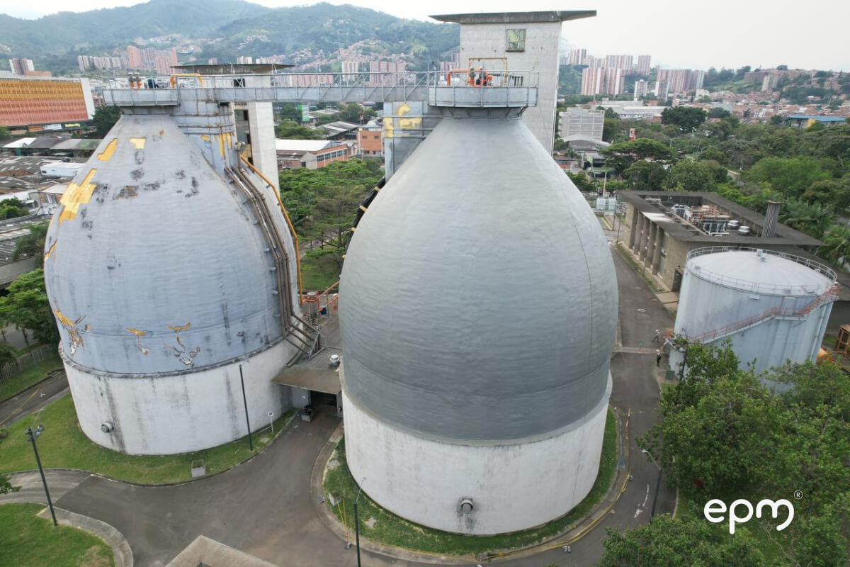 Vista aérea de biodigestores en planta de tratamiento, con áreas verdes y ciudad de fondo.