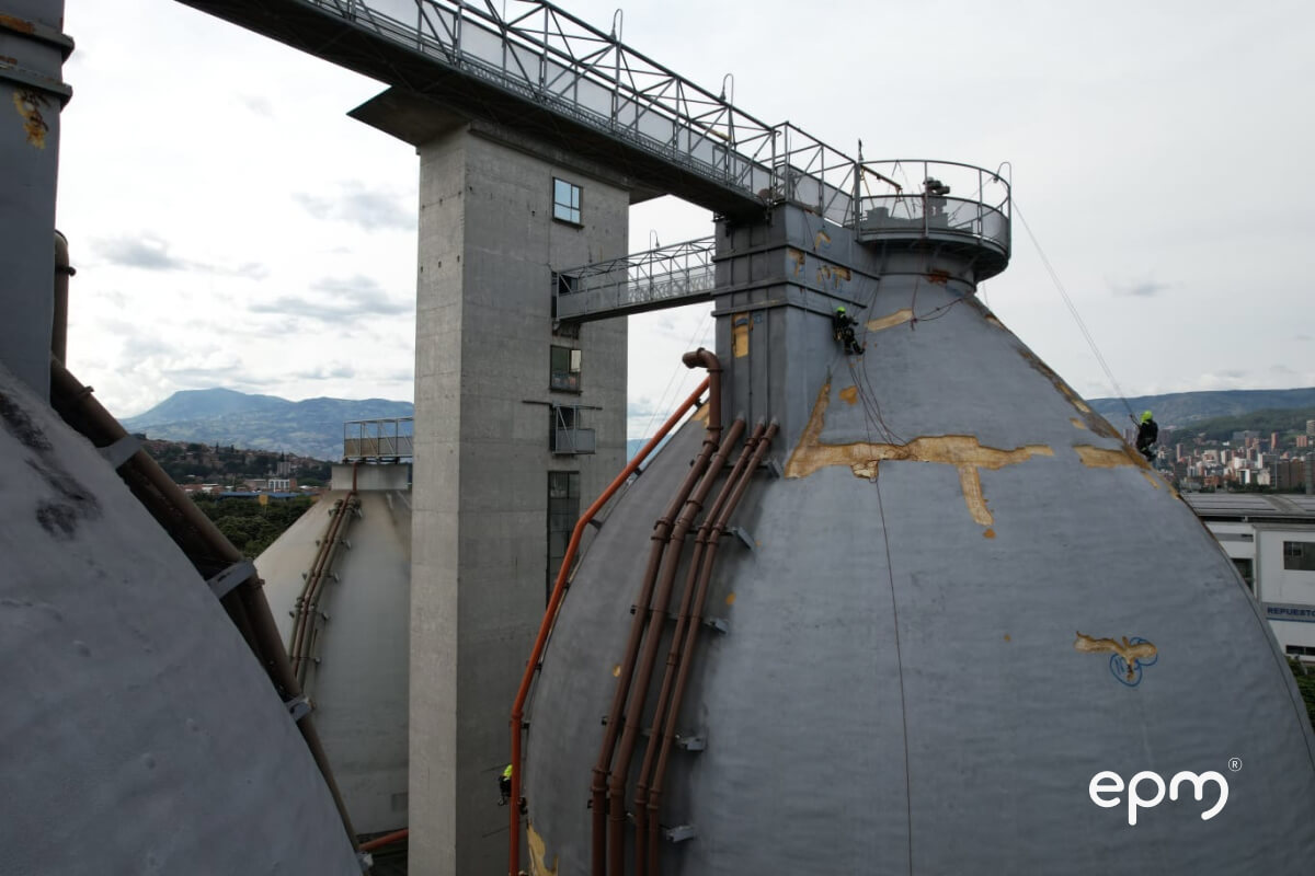 Operario de EPM en arnés trabajando en la cúpula de un biodigestor ovoide, con ciudad al fondo.