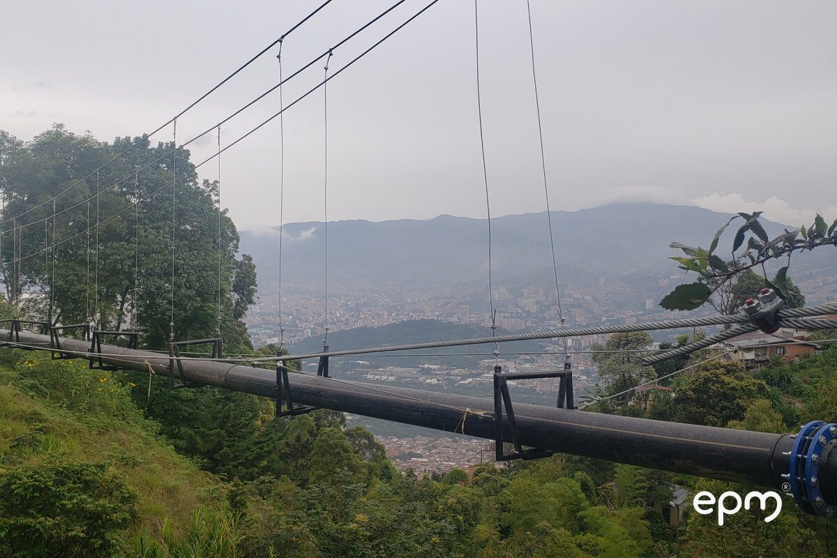 Fotografía de una tubería suspendida en el aire mediante cables de acero, con una vista panorámica de Medellín al fondo.