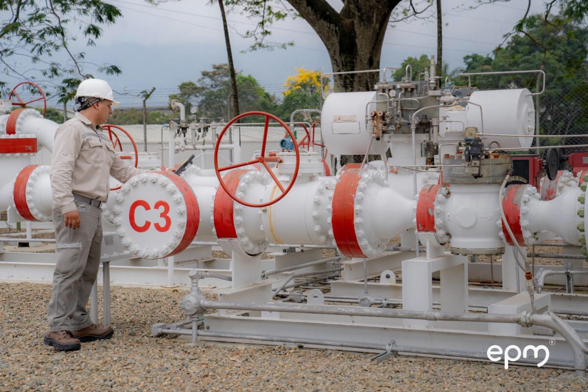 Fotografía de técnico de EPM con casco y uniforme inspecciona una estación de gas natural con tuberías blancas y rojas en una planta industrial.