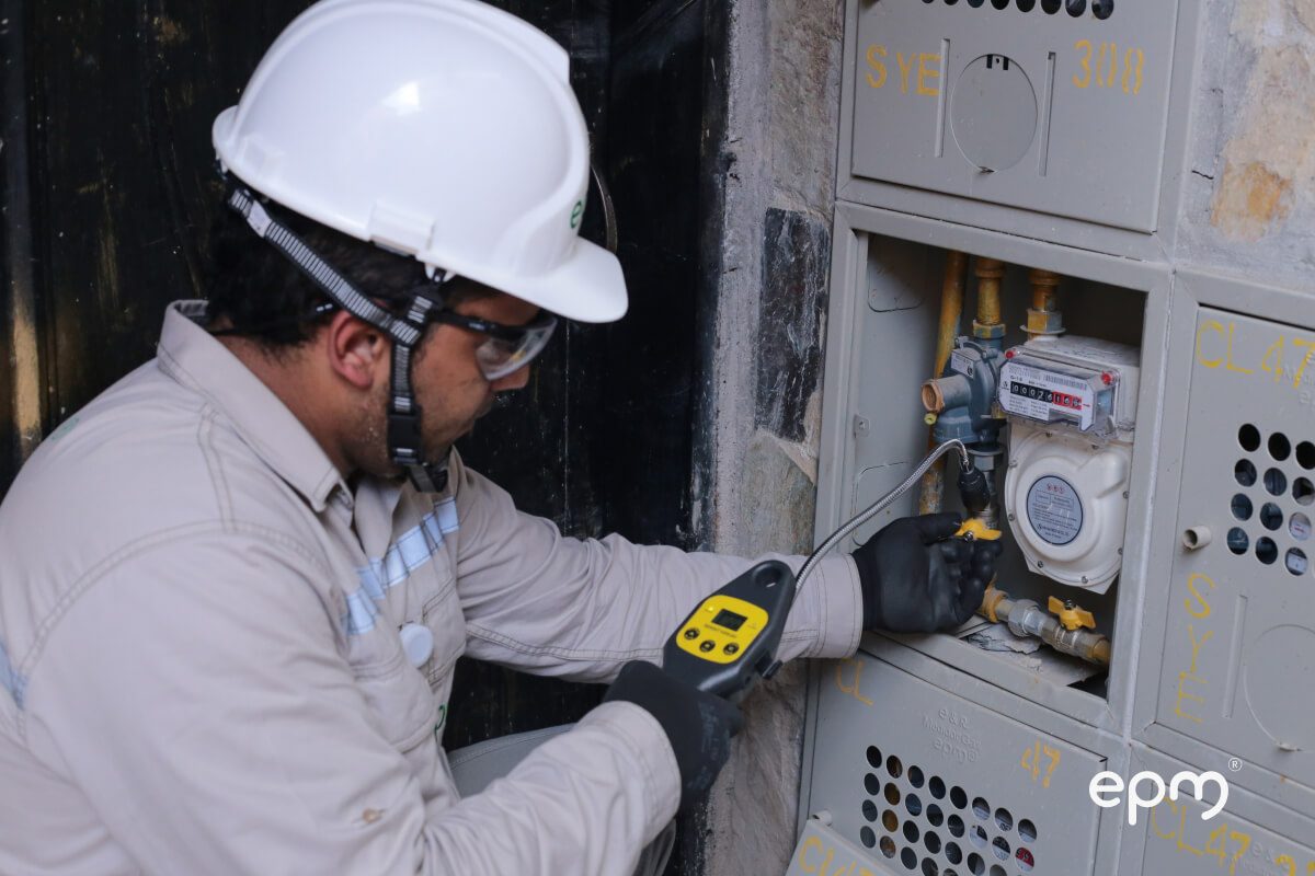 Fotografía de técnico de EPM con casco blanco y gafas de seguridad revisa y mide una conexión de gas en un medidor residencial.