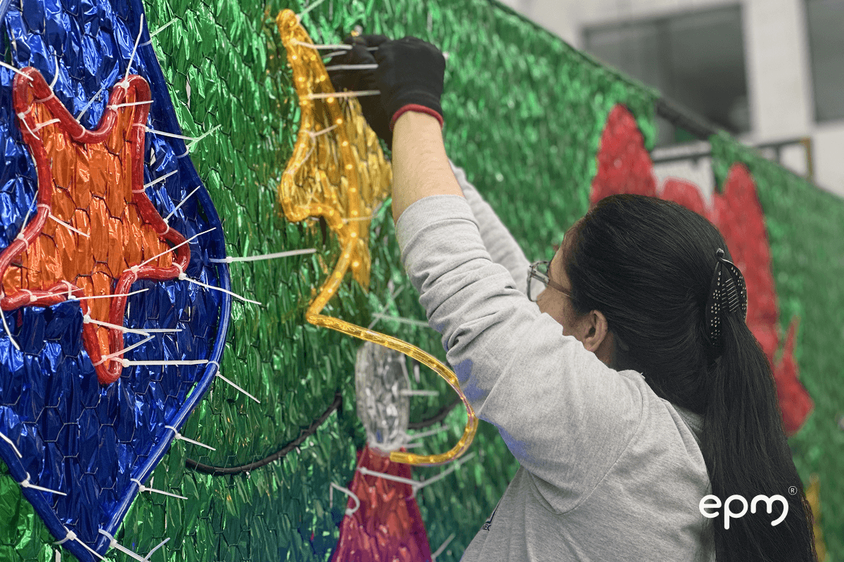 Imagen de mujer instalando luces navideñas en una estructura decorativa con fondo verde brillante.