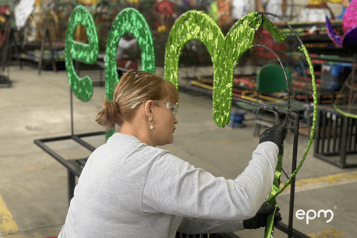 Fotografía de mujer en taller de montaje navideño, decorando la letra “m” del logo de EPM con papel metalizado verde.