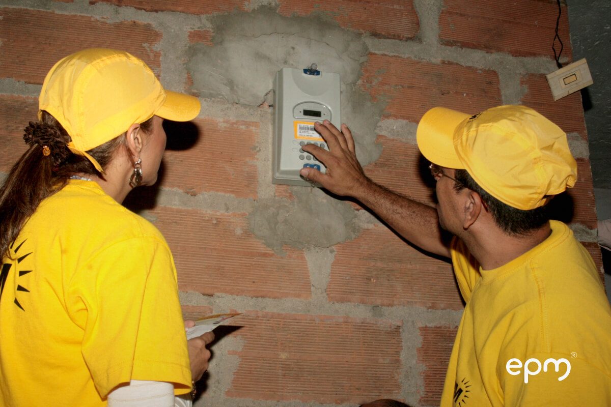Fotografía de dos trabajadores con uniforme amarillo de EPM instalan y configuran un medidor de energía en la pared de una vivienda.
