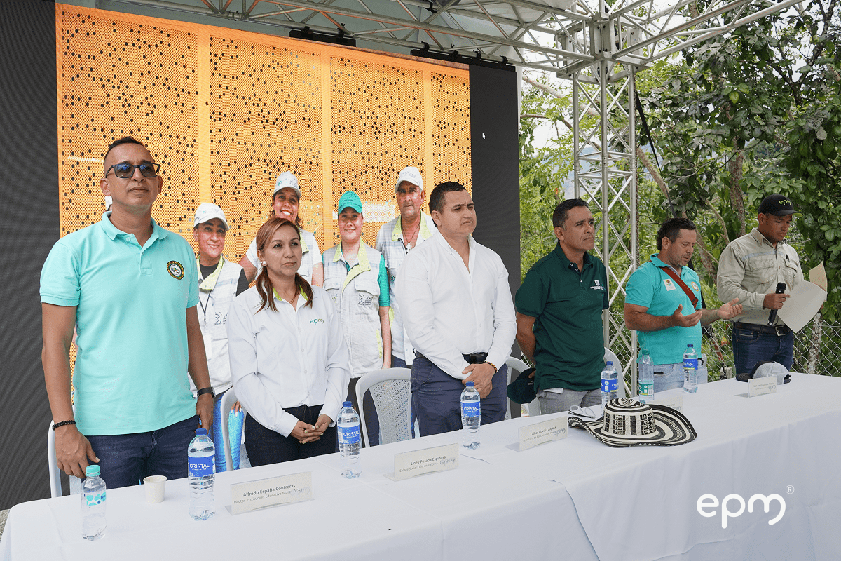 Personas de pie al frente de una mesa con los brazos cruzados. De fondo se observa pantalla y árboles