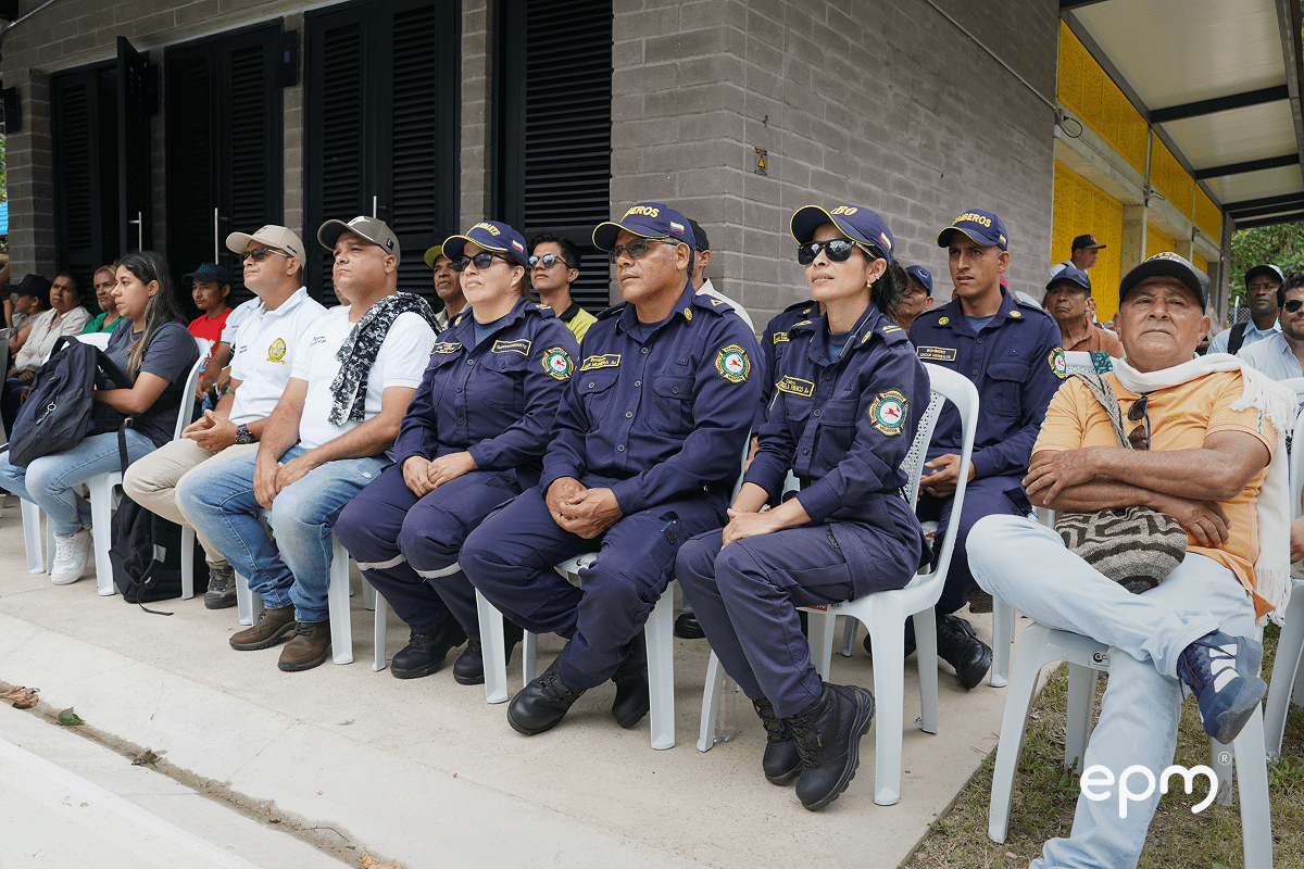 Personas reunidas sentadas mirando hacia el frente atentamente. De fondo se observan paredes en color gris y puertas negras