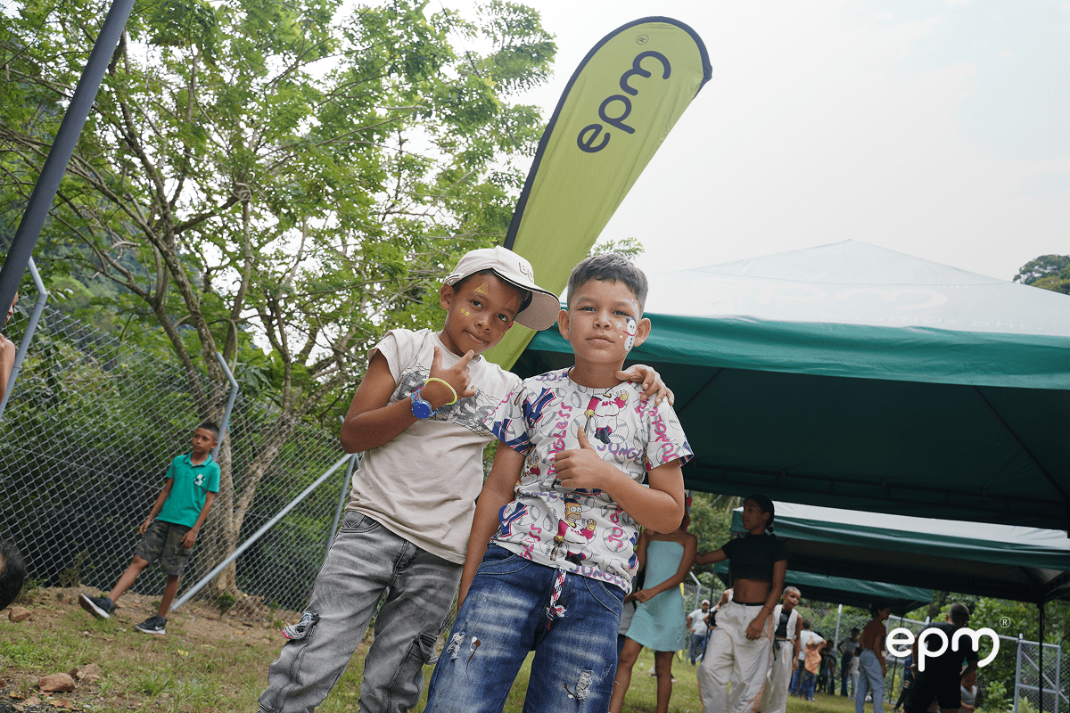Niños con la cara pintada abrazados y levantando una mano. De fondo se observa carpa en color verde, personas y árboles